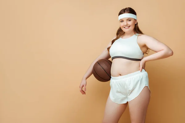 Plus size sportswoman with ball and hand on hip smiling and looking at camera on beige — Stock Photo
