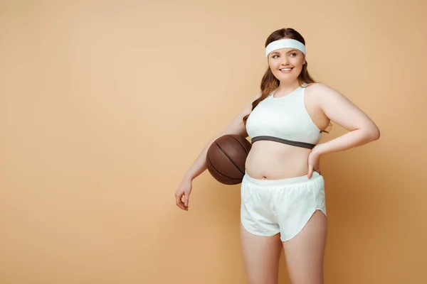 Deportista de talla grande con la mano en la cadera y la pelota sonriendo y mirando hacia otro lado en beige - foto de stock