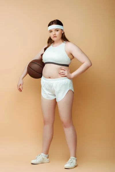 Plus size sportswoman with hand on hip and ball looking at camera on beige — Stock Photo
