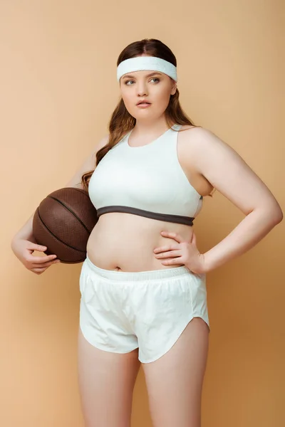 Plus size sportiva con mano sul fianco guardando altrove e tenendo la palla su sfondo beige — Foto stock