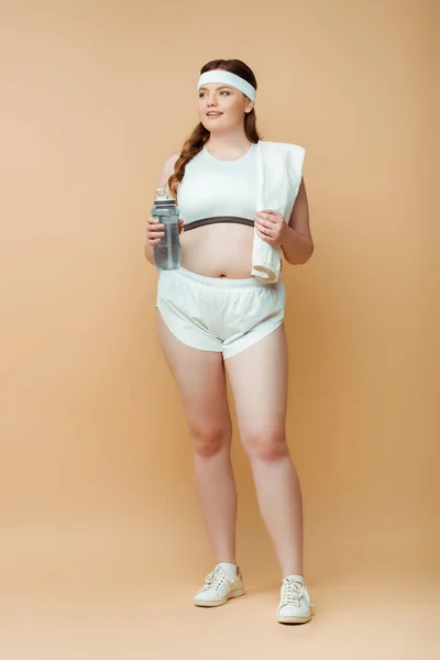 Plus size sportswoman with towel and sports bottle looking away on beige — Stock Photo