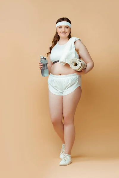 Plus size sportswoman with fitness mat and sports bottle looking away and smiling on beige — Stock Photo