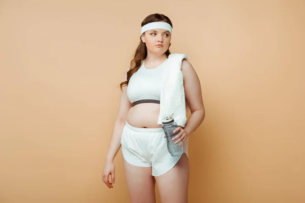 Plus size sportswoman with towel and sports bottle looking away on beige — Stock Photo