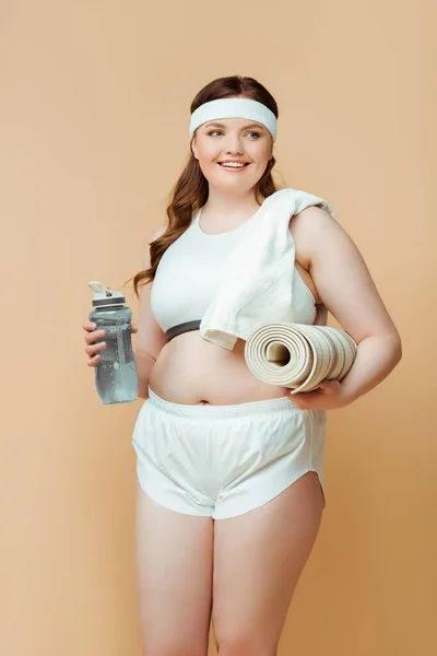 Além de mulher tamanho sorrindo, olhando para longe e segurando garrafa de esportes com tapete de fitness isolado no bege — Fotografia de Stock