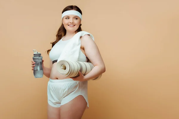 Grande taille femme souriant, regardant loin et tenant bouteille de sport avec tapis de fitness sur beige — Photo de stock