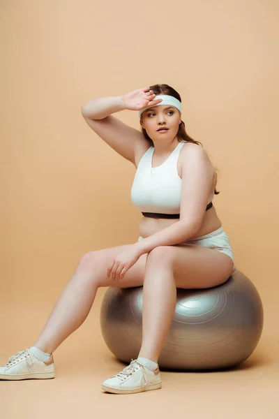 Plus size sportswoman raising hand and looking away on fitness ball on beige — Stock Photo