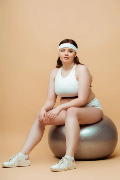 Plus size sportswoman looking at camera on fitness ball on beige — Stock Photo