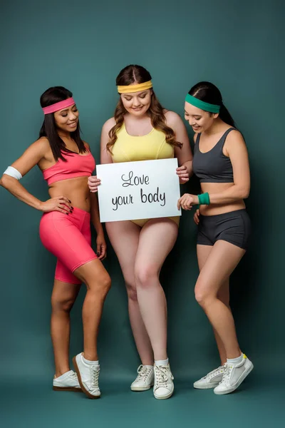 Sportive multietniche che sorridono e mostrano cartello con amore il tuo corpo lettering su sfondo verde — Foto stock