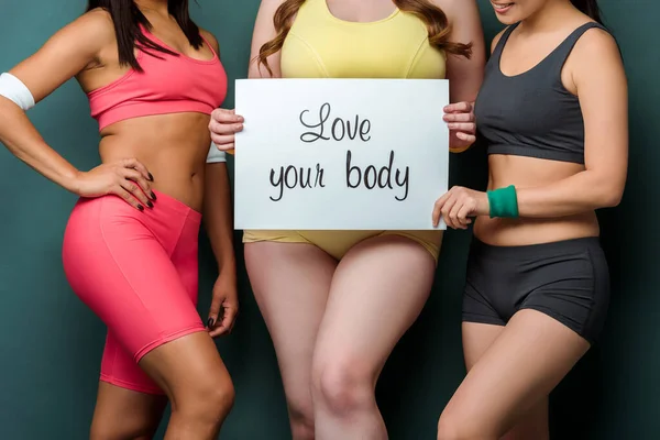 Cropped view of multicultural sportswomen showing placard with love your body lettering on green background — Stock Photo