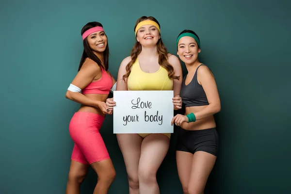 Sport multiculturali donne sorridenti, guardando la fotocamera e mostrando cartello con amore il tuo corpo lettering su sfondo verde — Foto stock