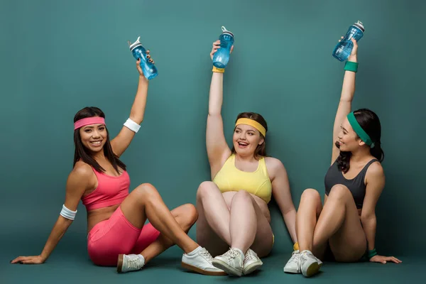 Desportistas multiétnicos com pernas cruzadas sorrindo e levantando as mãos com garrafas de esportes no fundo verde — Fotografia de Stock