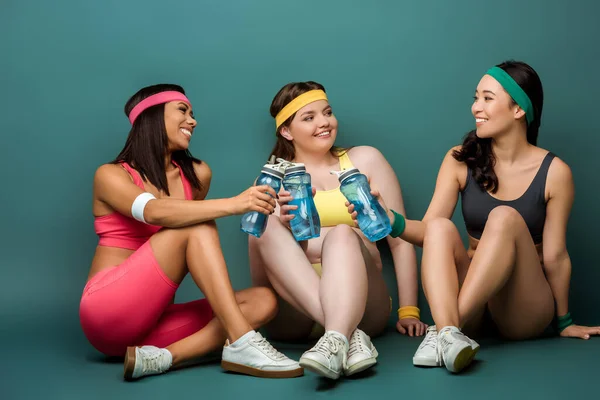 Sportives multiethniques assis avec les jambes croisées, souriant, tenant des bouteilles de sport et se regardant sur le vert — Photo de stock