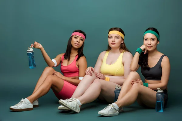 Multiethnische Sportlerinnen sitzen mit Sportflaschen und blicken in die Kamera auf Grün — Stockfoto