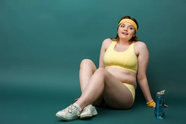 Plus-Size-Sportlerin sitzt mit überkreuzten Beinen neben Sportflasche auf grünem Hintergrund — Stockfoto