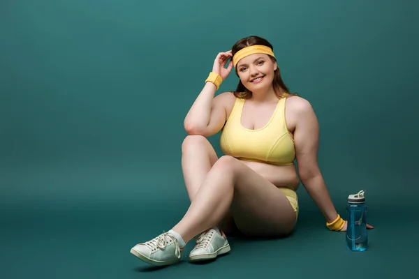 Plus-Size-Sportlerin mit überkreuzten Beinen lächelt und blickt in die Kamera neben Sportflasche auf Grün — Stockfoto