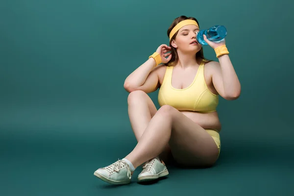 Plus-Size-Sportlerin sitzt mit überkreuzten Beinen und trinkt Wasser auf grünem Hintergrund — Stockfoto