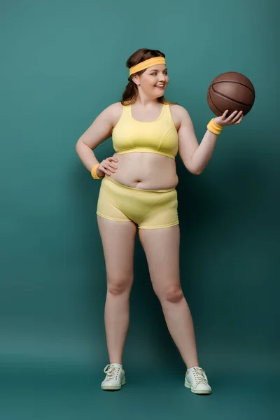 Plus size sportswoman with hand on hip looking at ball and smiling on green background — Stock Photo