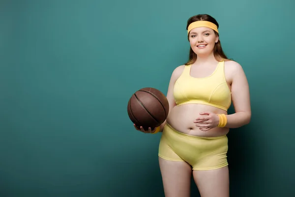 Plus-Size-Sportlerin mit Ball blickt in die Kamera und lächelt auf grünem Hintergrund — Stockfoto