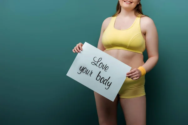 Cropped view of plus size sportswoman smiling and showing placard with love your body lettering on green — Stock Photo