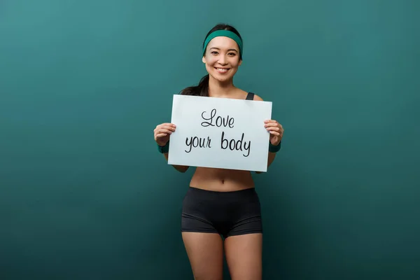 Asiática deportista mirando a la cámara, sonriendo y mostrando cartel con amor su cuerpo letras en verde - foto de stock