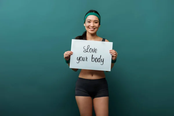 Asiatique sportive regardant la caméra, souriant et montrant la plaque avec amour votre corps lettrage sur vert — Photo de stock