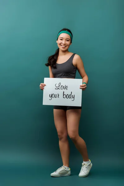 Attraente asiatica sportiva sorridente e tenendo cartello con amore il tuo corpo lettering su verde — Foto stock