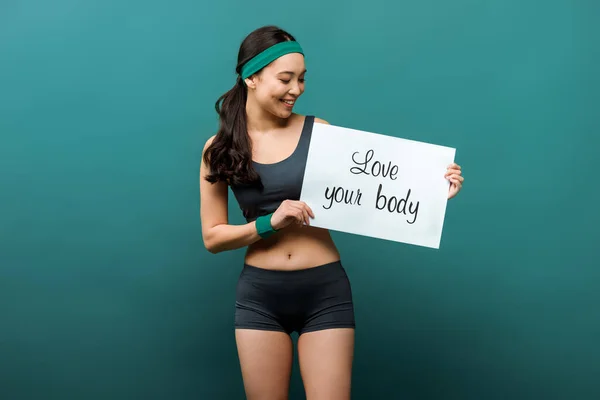 Front view of asian sportswoman smiling and showing placard with love your body lettering on green background — Stock Photo