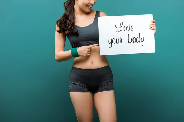 Cropped view of sportswoman smiling and pointing at placard with love your body lettering on green background — Stock Photo