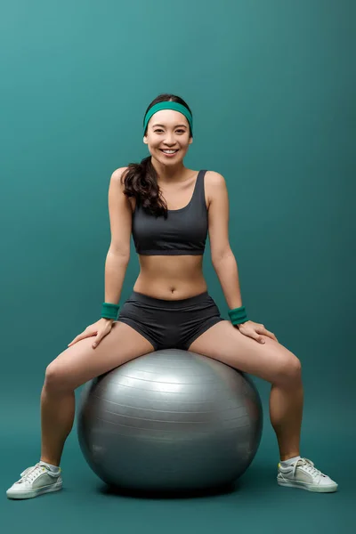 Asiatique sportive souriant et regardant la caméra sur la balle de fitness sur fond vert — Photo de stock