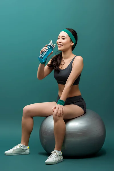 Asiática deportista sonriendo y bebiendo agua en fitness pelota en verde - foto de stock