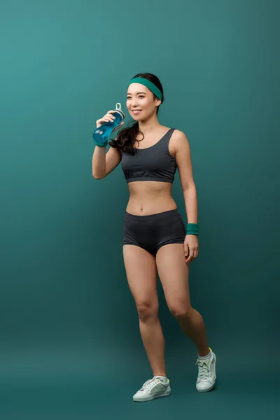 Asiática deportista con botella de deportes sonriendo sobre fondo verde - foto de stock