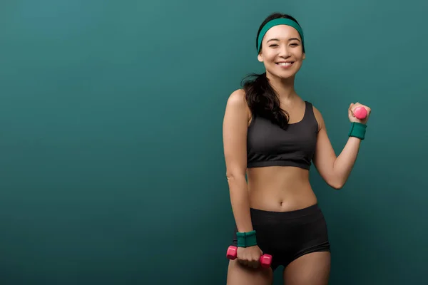 Asiática deportista sonriendo, mirando a la cámara y entrenando con mancuernas aisladas en verde - foto de stock