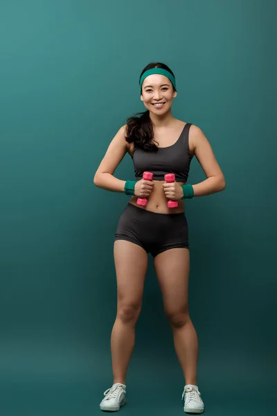 Vista frontal de la deportista asiática mirando a la cámara, sonriendo y sosteniendo pesas sobre fondo verde - foto de stock
