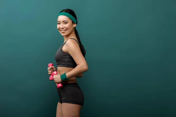 Vista lateral de una deportista asiática con mancuernas sonriendo y mirando a la cámara aislada en verde - foto de stock