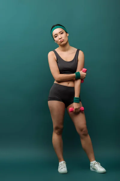 Втомилася азіатська спортсменка з гантелями, дивлячись на камеру на зеленому тлі — стокове фото