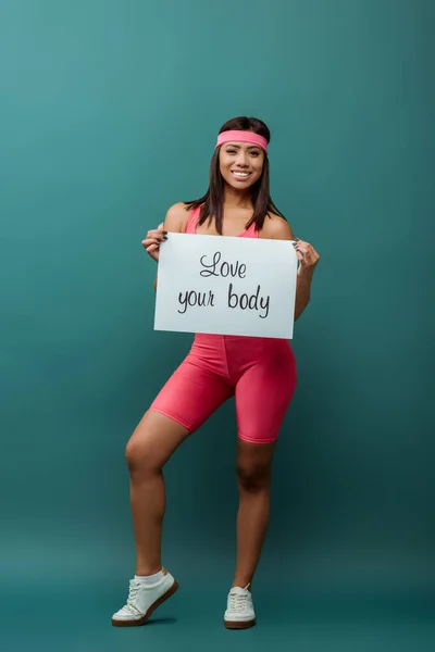 Felice sportswoman afro-americana sorridente, guardando la fotocamera e presentando cartello con amore la tua iscrizione del corpo sul verde — Foto stock