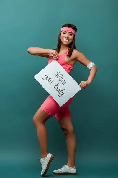 Happy african american sportswoman looking at camera and smiling with placard with love your body lettering on green background — Stock Photo