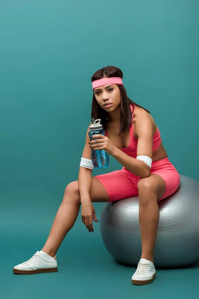 Atractiva deportista afroamericana sosteniendo botella deportiva y mirando a la cámara en la pelota de fitness sobre fondo verde - foto de stock