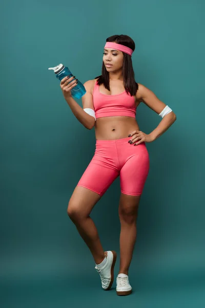 Femme sportive afro-américaine avec la main sur la hanche regardant la bouteille de sport sur fond vert — Photo de stock
