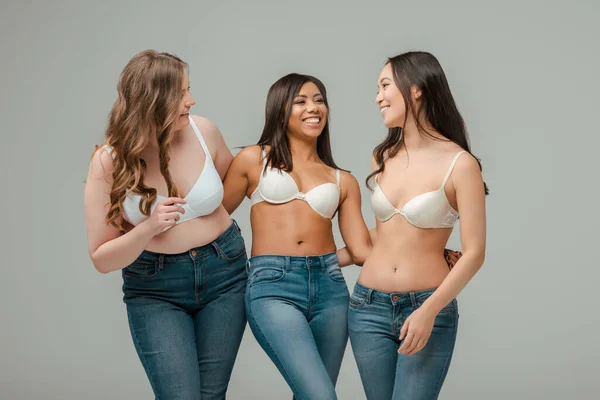 Donna multietnica in reggiseni che sorride, si abbraccia e si guarda isolata sul grigio — Foto stock