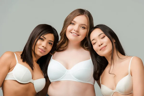 Donne multiculturali in reggiseni guardando la fotocamera e sorridendo isolate sul grigio — Foto stock