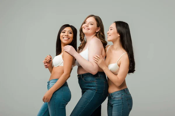 Felices mujeres multiculturales sonriendo y abrazándose aisladas en gris - foto de stock