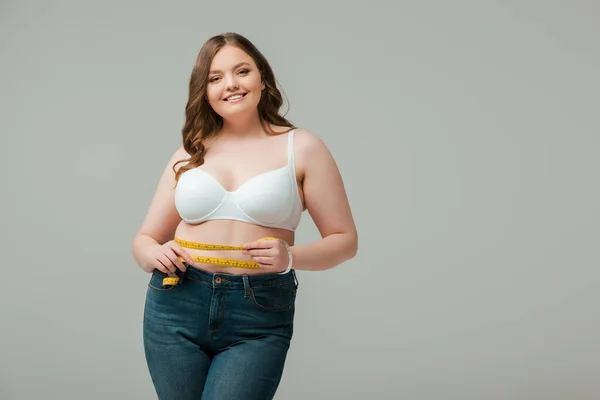 Plus size woman measuring waist, looking at camera and smiling isolated on grey — Stock Photo