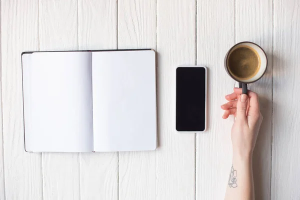 Vue recadrée de la main féminine avec une tasse de café près du smartphone et du portable sur fond en bois — Photo de stock