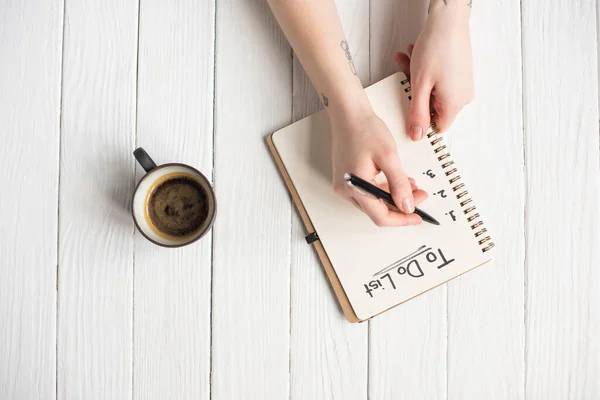 Vista cortada de mulher escrevendo em notebook com a lista de letras perto de xícara de café no fundo de madeira — Fotografia de Stock