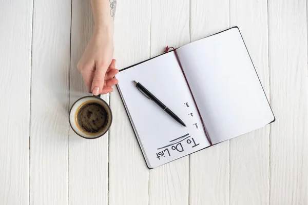 Vista cortada da mulher segurando xícara de café perto do caderno com letras da lista e caneta no fundo de madeira — Fotografia de Stock