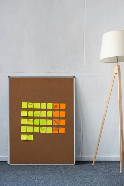Panneau marron avec des notes collantes jaunes et orange avec lampadaire près du mur — Photo de stock