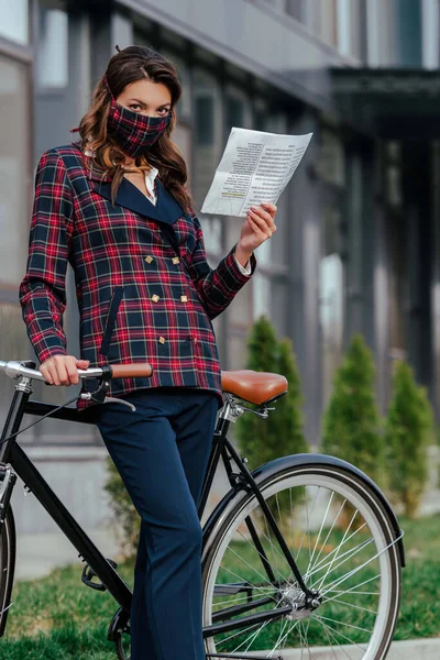 Businesswoman in plaid mask holding newspaper near bicycle — Stock Photo
