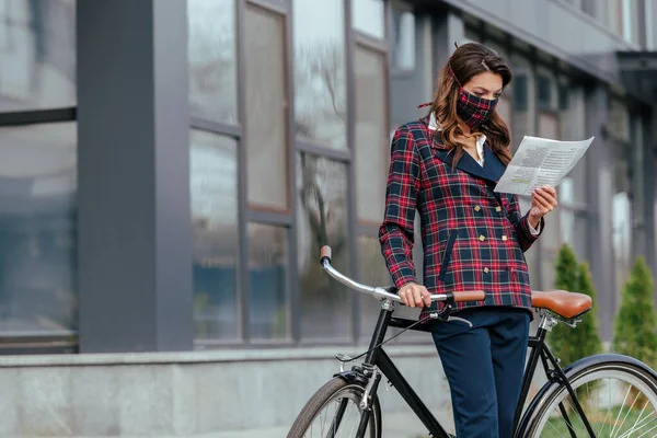 Femme d'affaires en masque à carreaux debout près de vélo et de lire le journal — Photo de stock