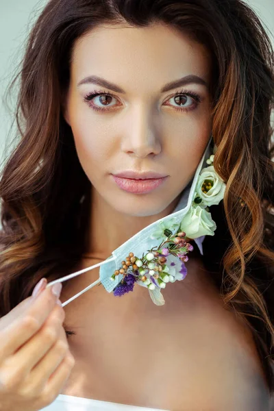 Приваблива жінка торкається маски для обличчя з квітами і дивиться на камеру ізольовано на сірому — стокове фото
